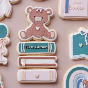 May include: A selection of decorated sugar biscuits on a pale pink surface. The biscuits feature a teddy bear, books with the text "First Edition", a balloon, a rainbow, and an open book with the text "Chapter One".