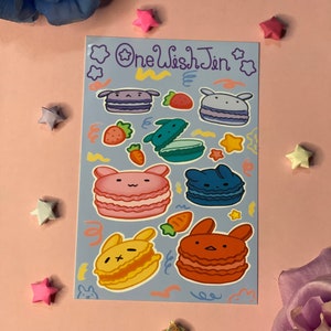 May include: A sheet of stickers featuring cute bunny macarons in various colors, including pink, blue, yellow, and red. The stickers are on a light blue background with white stars and swirls. The text "One Wish Jin" is printed in purple at the top of the sheet.