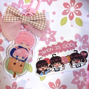 May include: An acrylic keychain featuring two stacked pink-haired characters and a brown paper bag, topped with a beige gingham and pink tulle bow. Beside it is a sticker with three dark-haired characters and the text TAMON IS GOD!