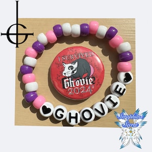 May include: A pink and purple beaded bracelet with a white heart charm and the words "I Survived Ghovie 2024" in black letters. The bracelet is next to a red button with a black and white cartoon rat and the words "I Survived Ghovie 2024".