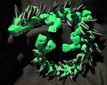 May include: A 3D-printed articulated dragon toy in bright green, black, and pink. The dragon has a detailed design with spikes and a segmented body, and is posed in a circular shape.