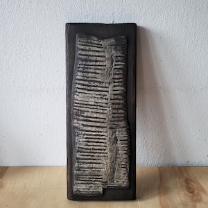 May include: A rectangular, dark-stained wooden wall art piece. The central design features a textured, light-colored, vertical form with horizontal lines, resembling a stylized landscape or abstract design. The artwork is mounted on a dark wooden frame.