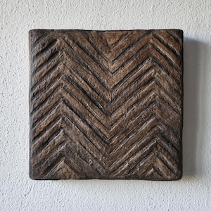 Può includere: Un'opera d'arte da parete decorativa quadrata, strutturata e marrone, con un motivo a spina di pesce. L'opera d'arte ha un aspetto rustico e invecchiato, con scanalature visibili e un colore marrone scuro.