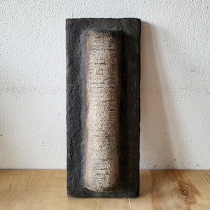 May include: A rectangular, textured wall art piece with a central, cylindrical design. The cylinder is light brown with vertical lines, set within a dark brown, recessed frame. The piece is displayed against a white wall and wooden surface.