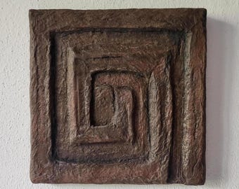 May include: Square, textured, brown wall art with a recessed spiral design. The artwork has a rough, earthy appearance and is mounted on a white wall. The design features concentric squares leading to a central point.