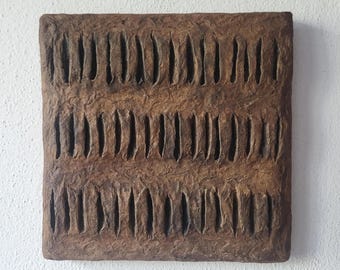 May include: A square, textured, brown artwork with a series of vertical, narrow indentations. The artwork has a rough, organic appearance, with a slightly uneven surface. The background is a plain white wall.