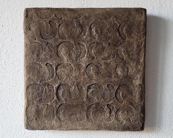 May include: A square, textured, brown wall art piece with a repeating pattern of curved shapes. The artwork is mounted on a white wall, providing a contrast to the earthy tones of the piece. The design is a series of connected, abstract forms.