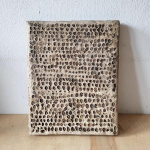 Könnte beinhalten: Ein rechteckiges Kunstwerk mit strukturierter Oberfläche und einem wabenartigen Muster. Die Oberfläche ist hellbeige mit zahlreichen dunkelbraunen, kreisförmigen Vertiefungen. Es wird vor einer weißen Wand und einer Holzoberfläche präsentiert.