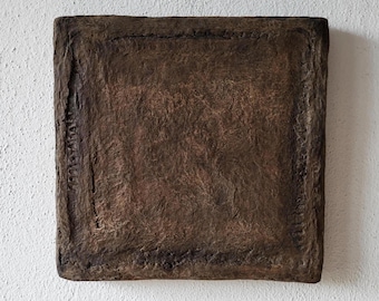 May include: A square, textured, brown artwork with a raised border. The artwork has a rough, uneven surface and is mounted on a white wall. The piece appears to be made of a natural material.