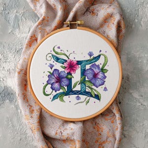 May include: A cross-stitch embroidery hoop featuring the Gemini zodiac symbol. The design includes a teal Gemini symbol, surrounded by purple and pink flowers and green leaves, set against a white fabric background. The wooden hoop is visible.