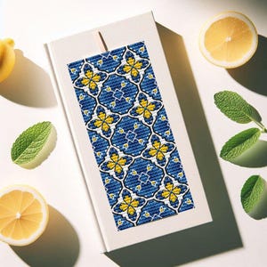 May include: A decorative bookmark with a blue and yellow floral pattern, resting on a closed white book. The bookmark features a repeating design of stylized flowers. Lemon slices and mint leaves are arranged around the book.