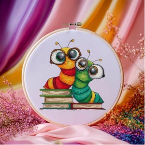 Può includere: Ricamo a punto croce in un telaio di legno con due bruchi in stile cartone animato con occhiali, seduti su libri. Un bruco è giallo e rosso, l'altro verde. Lo sfondo è un tessuto morbido e lilla chiaro.