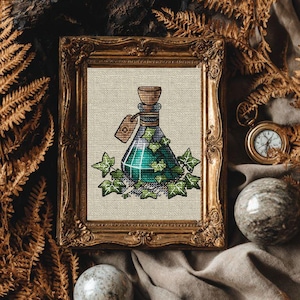 May include: Cross-stitch art of a green potion bottle with ivy, framed in a gold ornate frame. The bottle has a wooden stopper and a tag. The artwork is on a neutral linen background.