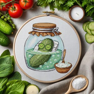 May include: A cross-stitch embroidery hoop featuring a cartoon pickle relaxing in a jar of brine. The pickle is green with closed eyes and a smile. A small wooden bowl of salt and a wooden spoon are also visible.