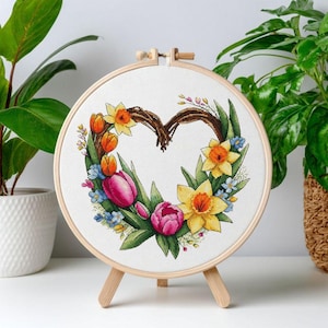 Puede incluir: Un bastidor de bordado circular de madera que contiene un patrón de punto de cruz de una corona en forma de corazón. El diseño presenta narcisos amarillos, tulipanes rosas y naranjas, y pequeñas flores azules con hojas verdes.
