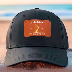 May include: Black baseball cap with a brown leather patch that says "galway" in gold lettering. The patch also features a gold harp and the text "EST. 1921" and "IRELAND".