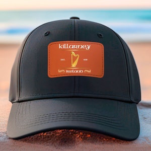 Puede incluir: Gorra de béisbol negra con un parche naranja que muestra el texto "killarney" y un arpa dorada con el texto "est. 810 ireland".