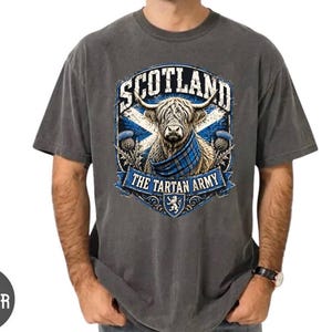 May include: A charcoal gray short-sleeve t-shirt featuring a graphic of a Highland cow wearing a blue tartan scarf. The design includes the text SCOTLAND and THE TARTAN ARMY, a blue and white saltire flag, thistle illustrations, and a lion crest.