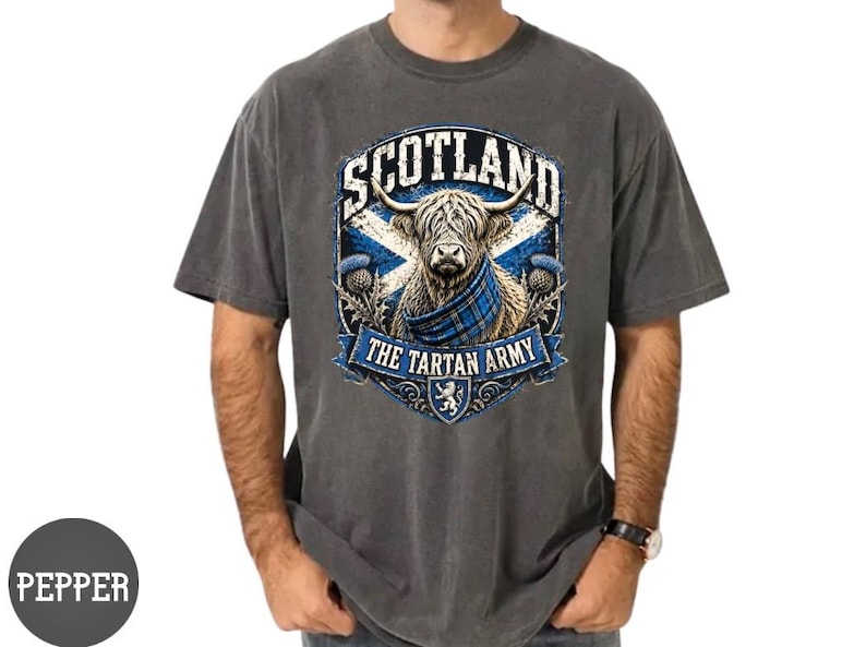 May include: A charcoal gray short-sleeve t-shirt featuring a graphic of a Highland cow wearing a blue tartan scarf. The design includes the text SCOTLAND and THE TARTAN ARMY, a blue and white saltire flag, thistle illustrations, and a lion crest.