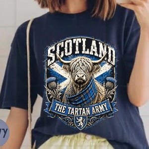 May include: A navy blue t-shirt featuring a graphic of a Highland cow wearing a blue plaid scarf. The design includes the text SCOTLAND and THE TARTAN ARMY, a blue and white saltire flag background, thistle illustrations, and a small lion crest.