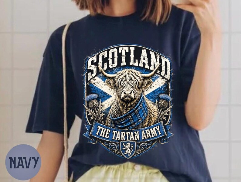May include: A navy blue t-shirt featuring a graphic of a Highland cow wearing a blue plaid scarf. The design includes the text SCOTLAND and THE TARTAN ARMY, a blue and white saltire flag background, thistle illustrations, and a small lion crest.