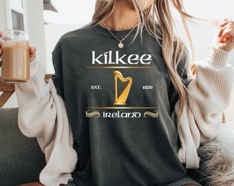 May include: Dark gray t-shirt with the word "kilkee" above a gold harp graphic and the word "Ireland". The shirt also has the text "EST. 1820". The person is holding a glass of a light brown beverage.