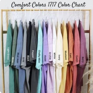 May include: A Comfort Colors 1717 color chart featuring a row of short-sleeve t-shirts hanging on a rack. The shirts are displayed in various colors including light green, moss, blue jean, gray, black, pepper, chalky mint, violet, orchid, white, ivory, mustard, yam, cumin, and berry.
