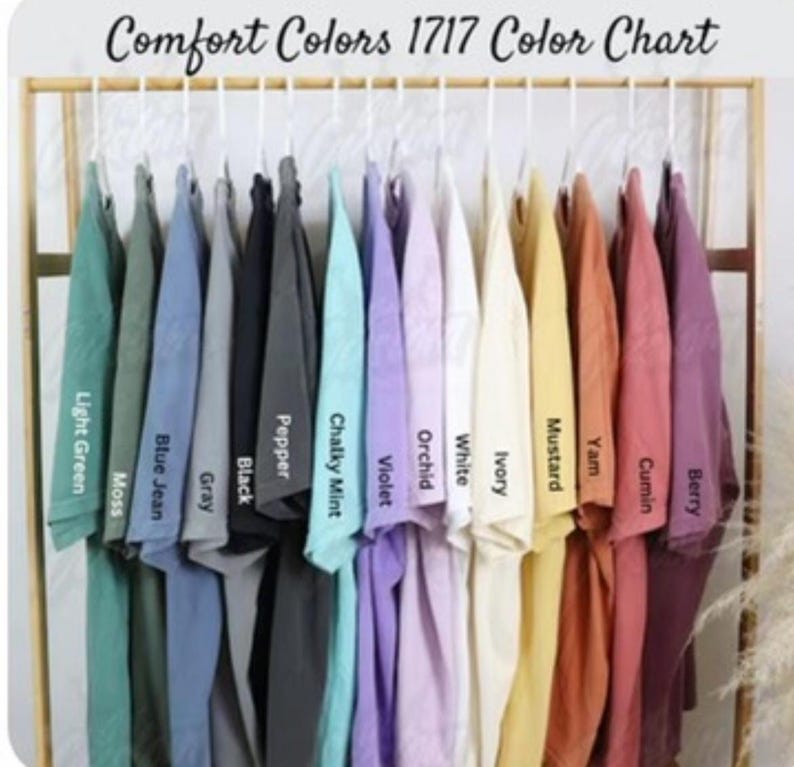 May include: A Comfort Colors 1717 color chart featuring a row of short-sleeve t-shirts hanging on a rack. The shirts are displayed in various colors including light green, moss, blue jean, gray, black, pepper, chalky mint, violet, orchid, white, ivory, mustard, yam, cumin, and berry.