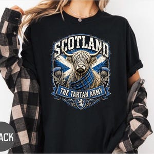 May include: A black t-shirt featuring a graphic print of a Highland cow wearing a blue plaid scarf. The design includes the text SCOTLAND and THE TARTAN ARMY, a blue and white saltire flag background, thistle illustrations, and a small lion crest.