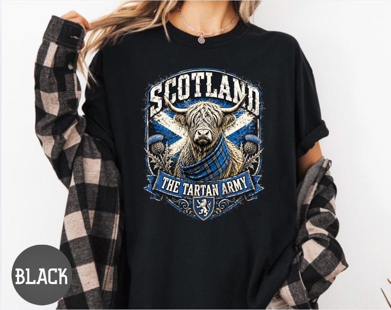 May include: A black t-shirt featuring a graphic print of a Highland cow wearing a blue plaid scarf. The design includes the text SCOTLAND and THE TARTAN ARMY, a blue and white saltire flag background, thistle illustrations, and a small lion crest.
