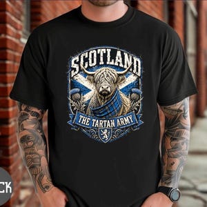 May include: A black short-sleeved t-shirt featuring a graphic print of a Highland cow wearing a blue tartan scarf. The design includes the text SCOTLAND and THE TARTAN ARMY, a blue and white saltire flag background, and thistle illustrations.