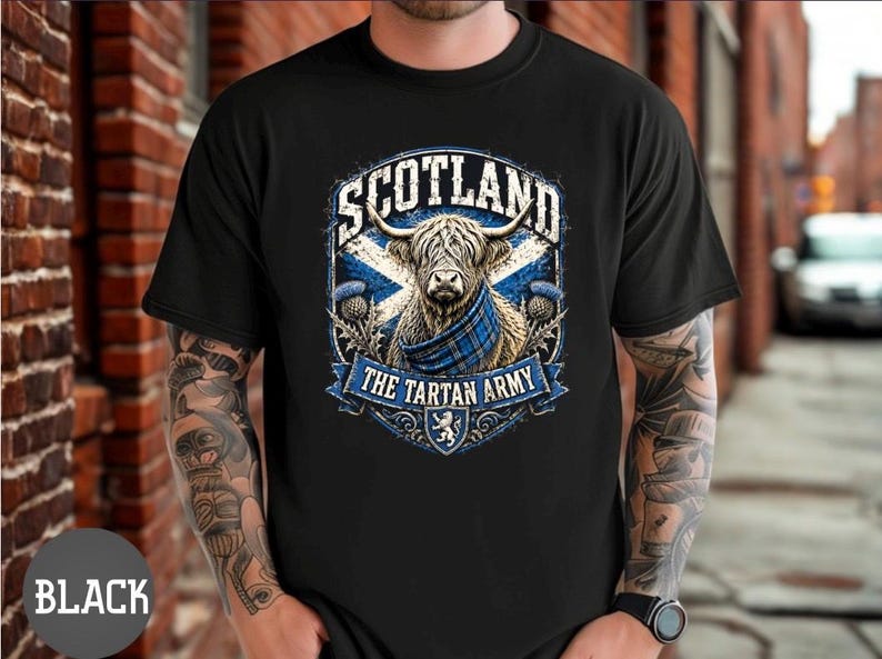 May include: A black short-sleeved t-shirt featuring a graphic print of a Highland cow wearing a blue tartan scarf. The design includes the text SCOTLAND and THE TARTAN ARMY, a blue and white saltire flag background, and thistle illustrations.