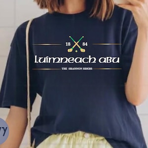 May include: A navy blue short-sleeve t-shirt featuring a graphic with two crossed green and yellow hurling sticks, the numbers 18 and 84, the text Luimneach Abu, and the text The Shannon Siders centered on the chest in white and gold lettering.