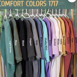 May include: A rack of 12 t-shirts in various colors, including light green, moss, black, pepper, gray, blue jean, chalky mint, violet, orchid, white, ivory, mustard, cumin, yam, and berry. The t-shirts are labeled with their color names. COMFORT COLORS 1717 is printed at the top of the image.