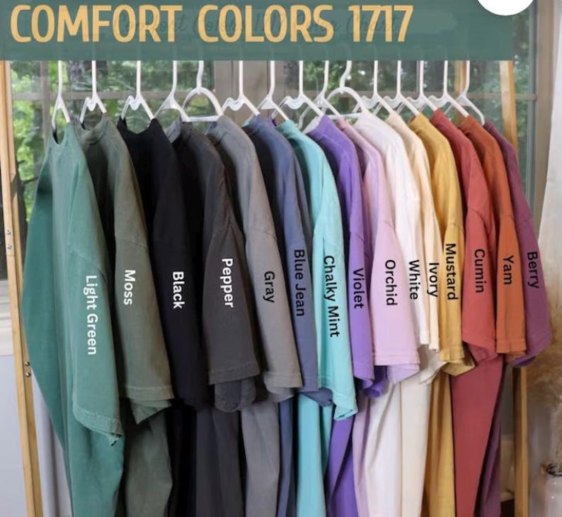 May include: A rack of 12 t-shirts in various colors, including light green, moss, black, pepper, gray, blue jean, chalky mint, violet, orchid, white, ivory, mustard, cumin, yam, and berry. The t-shirts are labeled with their color names. COMFORT COLORS 1717 is printed at the top of the image.