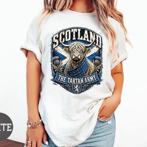 May include: A white short-sleeved t-shirt featuring a graphic of a Highland cow wearing a blue tartan scarf. The design includes the text SCOTLAND and THE TARTAN ARMY, set against a blue and white saltire flag background with thistle accents.