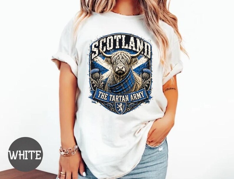 May include: A white short-sleeved t-shirt featuring a graphic of a Highland cow wearing a blue tartan scarf. The design includes the text SCOTLAND and THE TARTAN ARMY, set against a blue and white saltire flag background with thistle accents.