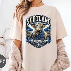 May include: An ivory short-sleeve t-shirt featuring a graphic of a Highland cow wearing a blue plaid scarf. The design includes the text SCOTLAND and THE TARTAN ARMY, a blue and white saltire flag, thistle illustrations, and a small lion emblem.