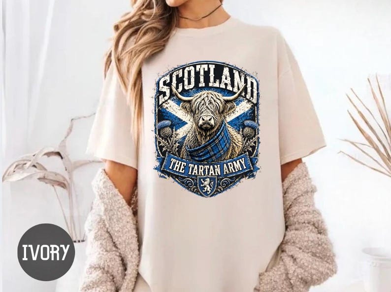 May include: An ivory short-sleeve t-shirt featuring a graphic of a Highland cow wearing a blue plaid scarf. The design includes the text SCOTLAND and THE TARTAN ARMY, a blue and white saltire flag, thistle illustrations, and a small lion emblem.