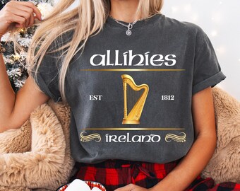 May include: Dark gray t-shirt with the word "allíbies" above a gold harp illustration. The text "EST 1812" and "íreland" are also displayed in gold. The shirt is a vintage style.