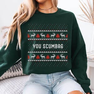 Puede incluir: Sudadera verde oscuro con un patrón festivo de renos y árboles de Navidad. Las palabras "YOU SCUMBAG" se muestran en letras blancas. Un diseño navideño humorístico.