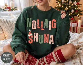 May include: A forest green crewneck sweatshirt featuring the text "NOLLAIG SHONA" in a distressed, multi-colored font. The letter "A" in the second line is topped with a small red and white Santa hat. The design has a textured, worn appearance.