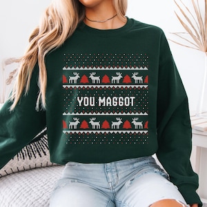 Puede incluir: Una sudadera verde oscuro con un patrón festivo con renos y árboles de Navidad en blanco y rojo. El texto "YOU MAGGOT" se muestra en letras blancas, creando un diseño navideño humorístico.
