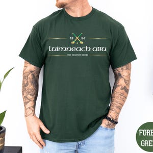 May include: A forest green short-sleeve t-shirt featuring a graphic with two crossed hurling sticks above the text 1884, the words Luimneach Abu, and the text The Shannon Siders, all centered on the chest in white and gold lettering.