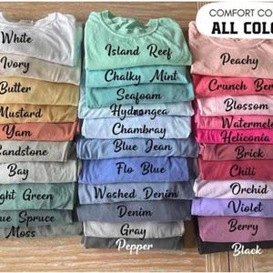 May include: A collection of folded t-shirts in various colors arranged in three vertical stacks. The text COMFORT COLORS ALL COLORS appears at the top right. The shirts are labeled with color names including White, Ivory, Butter, Mustard, Yam, Sandstone, Bay, Light Green, Blue Spruce, Moss, Island Reef, Chalky Mint, Seafoam, Hydrangea, Chambray, Blue Jean, Flo Blue, Washed Denim, Denim, Gray, Pepper, Peachy, Crunch Berry, Blossom, Watermelon, Heliconia, Brick, Chili, Orchid, Violet, Berry, and Black.