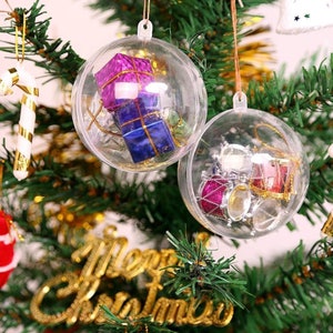May include: Two clear plastic Christmas baubles hanging on a Christmas tree. One bauble contains a small toy drum set, and the other contains small wrapped gifts. The baubles are hanging on a green Christmas tree with gold baubles and a gold "Merry Christmas" sign.