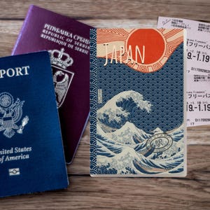 May include: A composition featuring a blue United States passport, a purple Serbian passport, and a Japanese travel journal. The journal displays a wave design with the word "JAPAN" and a red sun.