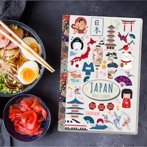 May include: A Japan travel journal with a light grey cover featuring colourful illustrations of Japanese cultural icons, including a sumo wrestler, pagoda, and lucky cat. The journal is next to a bowl of ramen and pickled ginger.