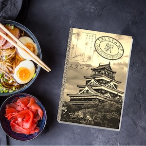 May include: A sepia-toned Eki Stamp Collection Book featuring a Japanese castle illustration. The book is open, displaying the cover design with Japanese text and a stamp-like emblem. A bowl of ramen with chopsticks and a small bowl of pickled ginger are also visible.
