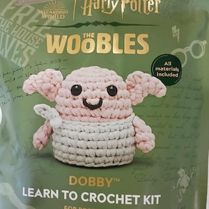 May include: A crochet kit featuring Dobby the house-elf from the Harry Potter series. The kit includes all materials needed to crochet a pink and white Dobby. The packaging is green with gold lettering and features the Wizarding World logo. The text on the packaging reads "Learn to Crochet Kit for Beginners".